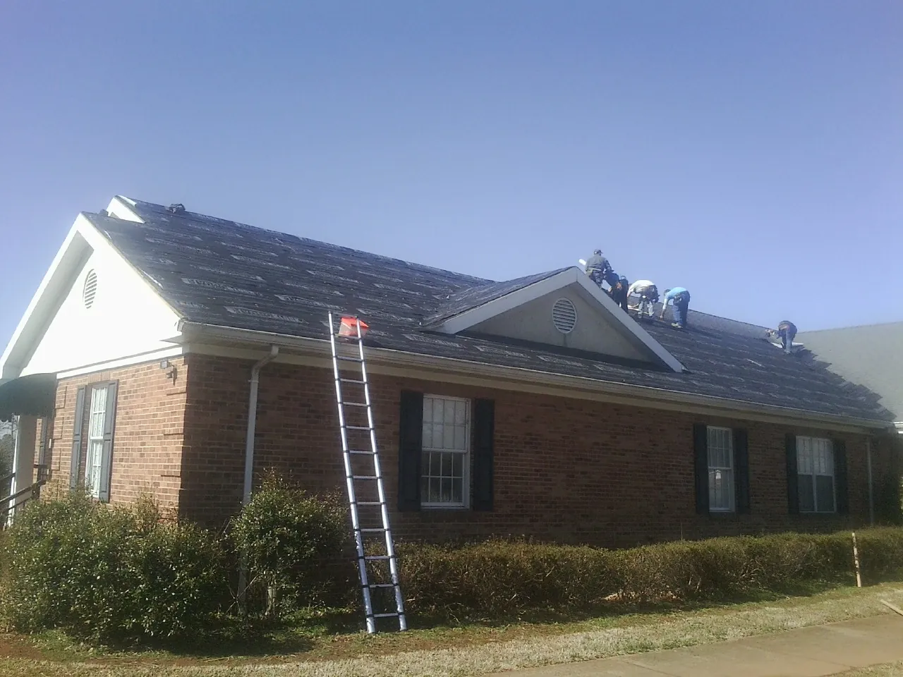 Crew tearing shingles off a brick ranch roof with a ladder set — Metal Roof Repair work by our crew in Highland